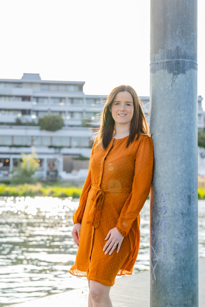 Braunhaarige Frau in orangenem Kleid lehnt lächelnd an einer grauen Säule